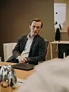 Mann i dress sitter ved møtebord og lytter mot en person utenfor bildekanten.