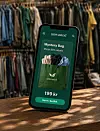 Smarttelefon med Unwasted-app som viser Mystery Bag-tilbud foran klesstativer i butikk.