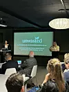 To presentatører viser Unwasted-logoen på lerret foran sittende publikum.