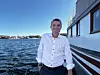 En mann i hvit skjorte står på et fartøy med vann og kystbebyggelse i bakgrunnen.