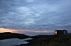 Kystlandskap med mørke svaberg, vann, skyer og en liten bygning til høyre.
