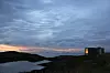 Kystlandskap med mørke svaberg, vann, skyer og en liten bygning til høyre.